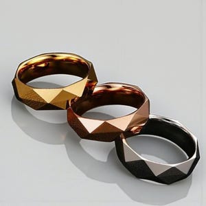 May include: Three geometric rings in gold, rose gold, and silver. Each ring features a faceted design, creating a play of light and shadow. The rings are displayed on a light-colored surface, showcasing their metallic sheen.