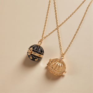 Puede incluir: Dos collares dorados con colgantes. Uno es un orbe negro y dorado con detalles intrincados. El otro es un colgante de orbe calado dorado con una pequeña gema. Ambos collares tienen delicadas cadenas doradas.