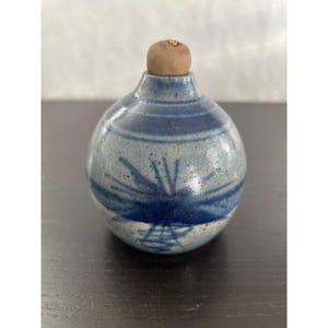 May include: A small, round ceramic bottle with a cork stopper. The bottle is primarily light blue with darker blue horizontal stripes and a stylized sunburst design. The bottle is approximately 4 inches tall.
