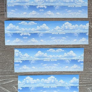 May include: Four rectangular stickers featuring a blue sky with fluffy white clouds. The stickers are arranged on a wooden surface. The sky design is consistent across all stickers, creating a cohesive aesthetic. The stickers are likely for crafting or decorative purposes.