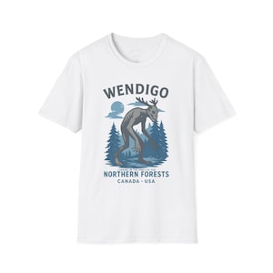 Può includere: T-shirt bianca con un'illustrazione grigia di un Wendigo, creatura con corna, in un ambiente forestale. Il testo "WENDIGO" è sopra l'illustrazione, con "LURKING IN THE PINES OF THE NORTHERN FORESTS CANADA USA" sotto.