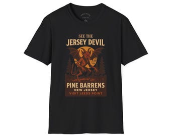 May include: Black t-shirt featuring a graphic of the Jersey Devil with text that reads "See the Jersey Devil, Pine Barrens, New Jersey, Visit Leeds Point." The graphic is in shades of brown, orange, and yellow.