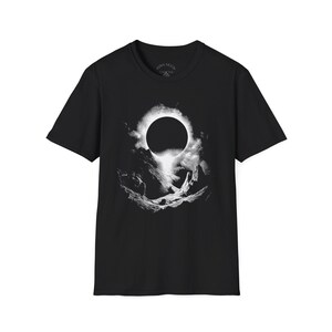 Puede incluir: Camiseta negra con un gráfico blanco que representa un eclipse solar sobre un paisaje montañoso. El diseño incluye un círculo oscuro rodeado de nubes y luz brillante, con el texto "KIRIN MOON" en el cuello.