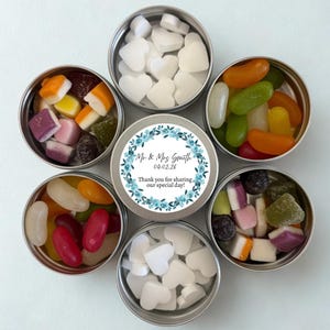 Pode incluir: Uma coleção de pequenas latas redondas prateadas cheias de doces coloridos. Os doces incluem doces em forma de coração e vários doces de gelatina. Uma lata central apresenta um design floral com o texto "Mr & Mrs Smith 04.02.26 Obrigado por partilhar o nosso dia especial!"