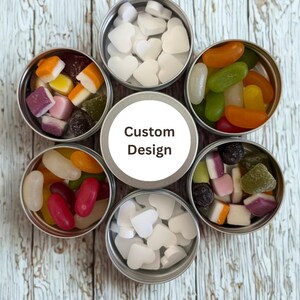 Pode incluir: Variedade de doces em pequenas latas redondas de metal dispostas em círculo. Os doces incluem doces brancos em forma de coração, jujubas e outras guloseimas coloridas. A lata central tem a inscrição "Custom Design".