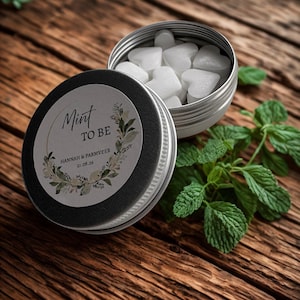 May include: A small, open silver tin filled with heart-shaped mints, next to a closed tin with the text "Mint to Be" and floral accents. Fresh mint leaves are scattered on a wooden surface.