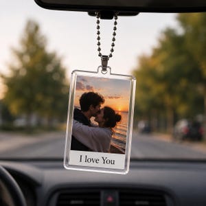 May include: A rectangular clear acrylic car air freshener or hanging ornament featuring a photograph of a couple embracing at sunset by the water. Below the image, a white rectangular section displays the black printed text I love You.