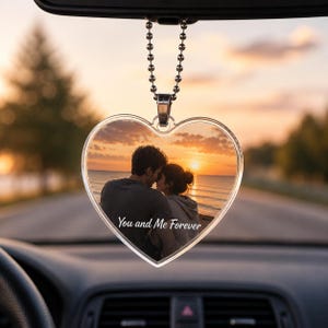 May include: A heart-shaped acrylic car ornament hanging from a silver ball chain. The ornament features a printed photograph of two people embracing at sunset by the ocean, with the white cursive text "You and Me Forever" printed below them.