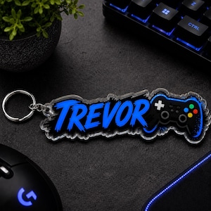 May include: A clear acrylic keychain featuring the name Trevor in a bold, bright blue brush-stroke font, accompanied by a black video game controller graphic with colorful buttons, attached to a silver metal split ring for keys.