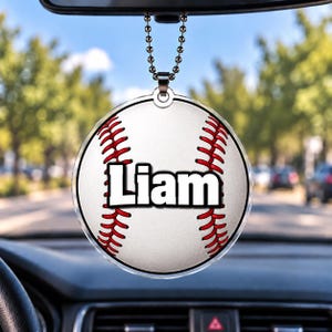 May include: A round, clear acrylic car ornament featuring a white baseball graphic with red stitching and the name Liam printed in bold, black-outlined white letters. The ornament hangs from a silver-toned metal ball chain.
