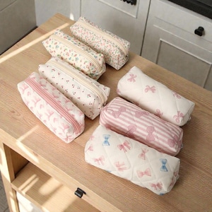 Puede incluir: Una colección de estuches para lápices con varios diseños florales y de lazos. Los estuches son rectangulares con cierre de cremallera y están hechos de tela. Los colores incluyen rosa, blanco y crema. Los estuches están colocados sobre una superficie de madera clara.