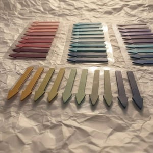 Pode incluir: Uma coleção de marcadores de página adesivos em forma de seta dispostos em folhas de suporte de plástico transparente. Os marcadores apresentam um gradiente de cores incluindo tons de rosa, vermelho, azul, roxo, amarelo e verde, apontando para a esquerda.
