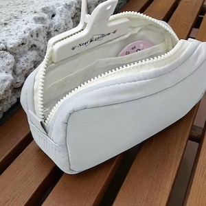 Op de afbeelding: Een lichtgrijze rechthoekige etui met een witte rits, gedeeltelijk open. Binnenin is een witte hanger met de tekst "Near Uniform" zichtbaar. Een klein roze cirkelvormig item bevindt zich ook in de etui. De etui ligt op een houten oppervlak.