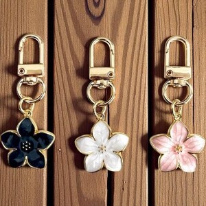 Puede incluir: Tres llaveros dorados con colgantes de flores en negro, blanco y rosa. Cada llavero tiene un cierre de mosquetón y un charm en forma de flor con ribete dorado. El fondo es una superficie de madera.