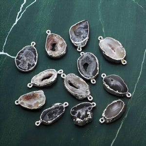May include: A selection of agate gemstone pendants with silver-coloured metal edging. The pendants are in various shapes and sizes, displaying a range of colours such as grey, brown, and white. Each pendant has a small loop for attaching to a chain or other jewellery component.