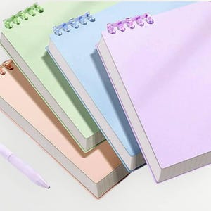 May include: Four pastel-coloured spiral-bound notebooks in pink, green, blue, and purple. Each notebook has a matching spiral binding and white paper. A light purple pen is in the foreground.