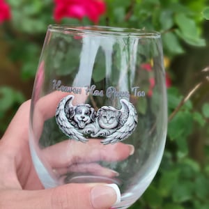 May include: A clear stemless wine glass with the text "Heaven Has Paws Too." The glass features an illustration of a dog and a cat with angel wings. The background is blurred with red flowers and green foliage.