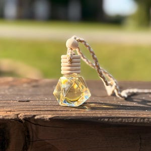 May include: A small, faceted glass bottle with a wooden cap and a braided rope. The bottle contains a yellow liquid and is designed to hang. The background is blurred, with a wooden surface.