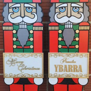 May include: Two wooden Christmas nutcracker figures. Each figure is painted with red, green, and gold accents. One figure has "Merry Christmas" on a sign, the other has "Ybarra".