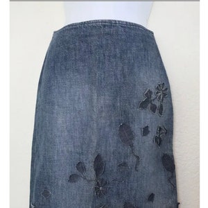 May include: A dark blue denim skirt with a floral design cut out of the fabric. The skirt is knee-length and has a straight silhouette.