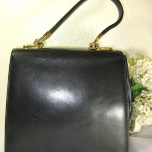 May include: A black leather handbag with a gold-tone handle. The bag has a rectangular shape and a smooth, polished finish.