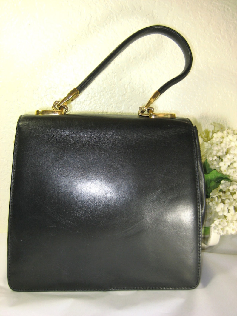 May include: A black leather handbag with a gold-tone handle. The bag has a rectangular shape and a smooth, polished finish.
