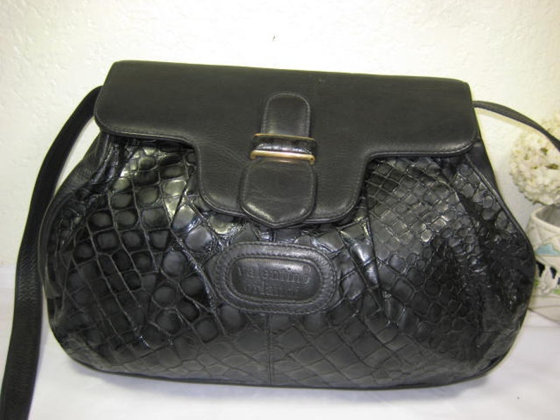 May include: A black leather handbag with a crocodile-embossed pattern and a gold-toned clasp. The bag has a single strap and a flap closure.