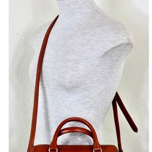 Puede incluir: Un bolso de mano de cuero marr&oacute;n con una hebilla dorada y una correa larga ajustable. El bolso tiene un asa superior y una correa para el hombro.