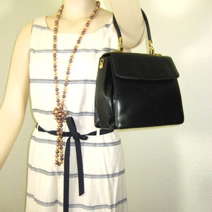 May include: A black leather handbag with a gold clasp and a gold chain strap. The bag is being held by a mannequin wearing a white and black striped dress with a brown and gold beaded necklace.