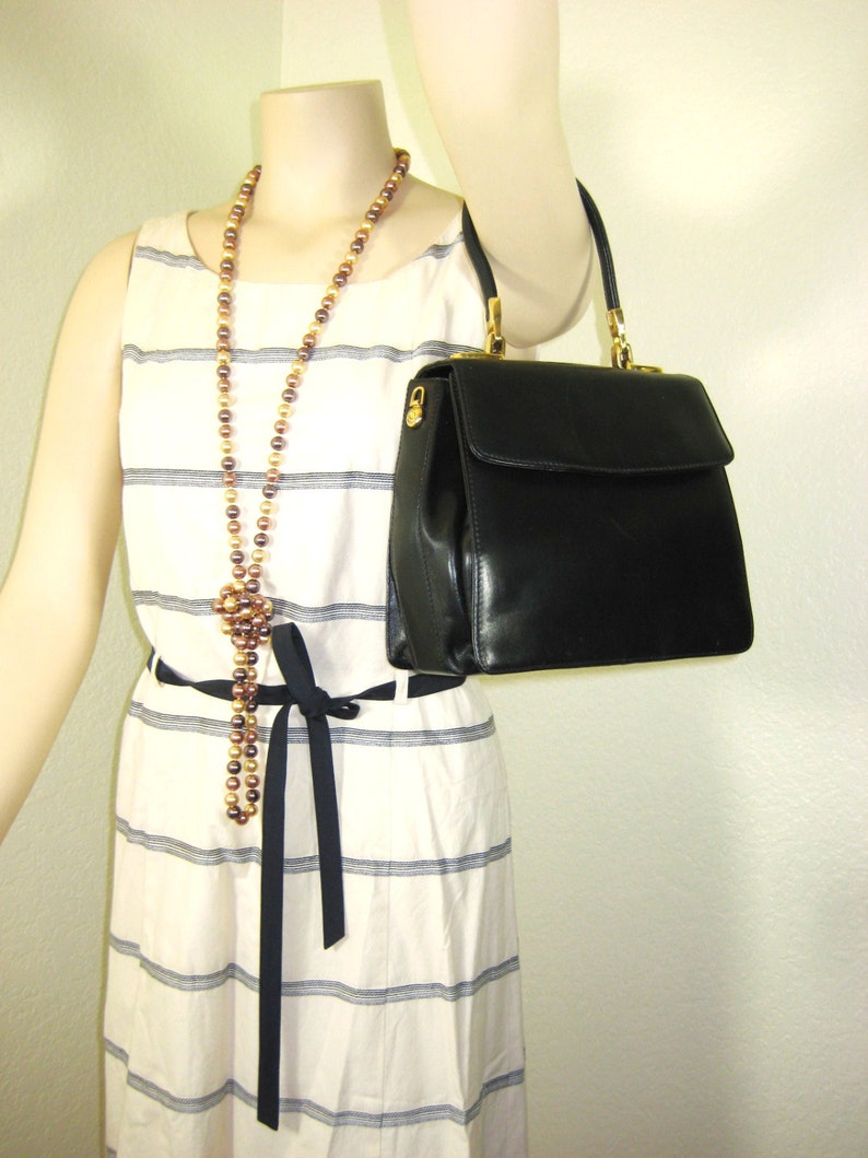 May include: A black leather handbag with a gold clasp and a gold chain strap. The bag is being held by a mannequin wearing a white and black striped dress with a brown and gold beaded necklace.