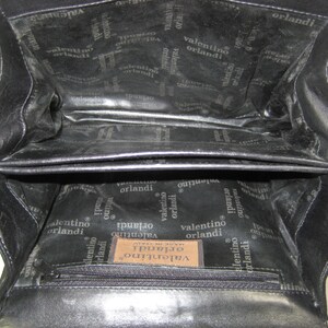 May include: Black leather handbag with a zippered pocket and a label that reads "Valentino Orlandi Made in Italy". The inside of the bag is lined with a black fabric with the Valentino Orlandi logo repeated throughout.