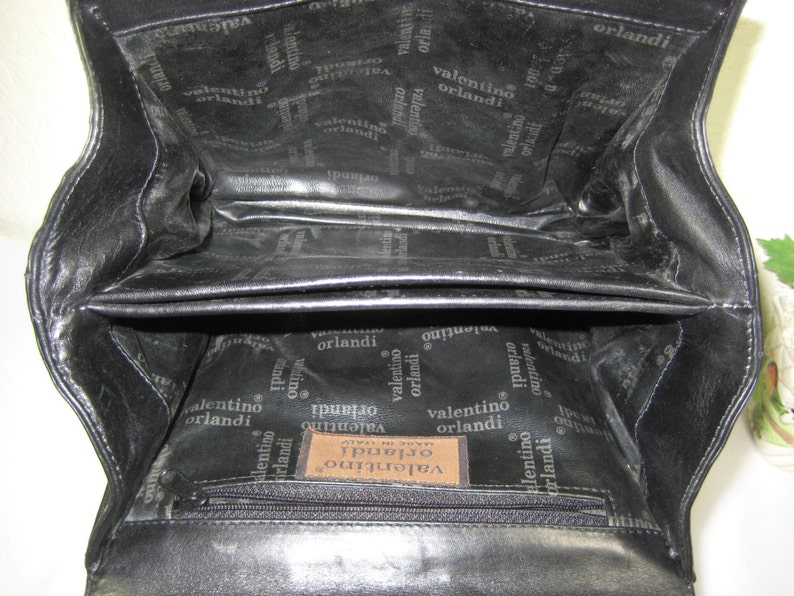 May include: Black leather handbag with a zippered pocket and a label that reads "Valentino Orlandi Made in Italy". The inside of the bag is lined with a black fabric with the Valentino Orlandi logo repeated throughout.