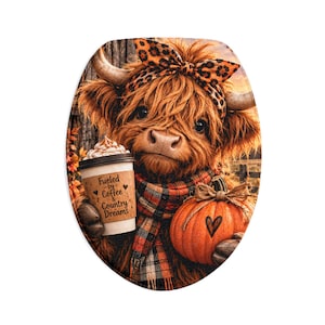 Op de afbeelding: Een ovale toiletbrilhoes met een pluizige bruine Schotse hooglander met een hoofdband met luipaardprint en een geruite sjaal. De koe houdt een koffiebeker vast met de tekst Fueled by Coffee & Country Dreams en een oranje pompoen met een hartje.