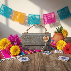May include: Gray leather handbag with floral embossing, a brown handle, and a decorative tassel. The bag is adorned with a wooden heart charm that says "I Love Artisanal". The scene includes colorful flowers, coasters, and a festive banner.