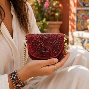 May include: A small, deep red, semi-circular handbag featuring an intricate, embossed floral pattern across its front flap. The bag is equipped with a polished gold-tone metal chain strap that attaches to the sides with matching gold-tone lobster clasps.