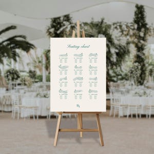 Op de afbeelding: Een crèmekleurige plattegrond op een houten ezel. De kaart toont een golfbaanontwerp met tafelnummers en -namen in een blauwgroene kleur. De woorden "Seating chart" staan bovenaan. De initialen "HA" staan onderaan.