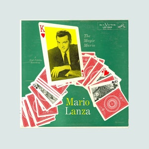 Pode incluir: Uma capa de álbum de vinil verde com um design de cartas de jogo vermelhas. A capa do álbum apresenta uma foto de Mario Lanza, um cantor, em uma moldura amarela. O título do álbum é "The Magic Mario" e o selo do disco é RCA Victor LM-1943.