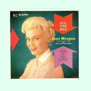 May include: A vintage vinyl record cover featuring a woman with blonde hair and a pink and white striped shirt. The cover is red, green, and pink with the title "All The Way" and the artist "Jane Morgan, The Fascination Girl and The Troubadors".