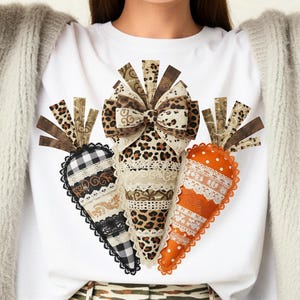Puede incluir: Camiseta blanca con tres diseños decorativos de zanahorias. Cada zanahoria presenta un patrón único, incluyendo estampado de leopardo, cuadros vichy y naranja con lunares blancos. Las zanahorias están adornadas con encaje y cintas, creando un aspecto festivo y caprichoso.