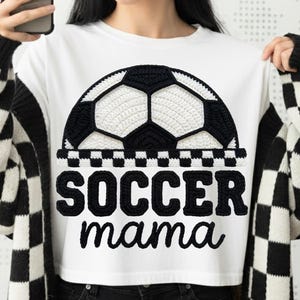 May include: White t-shirt featuring a black and white crocheted soccer ball design. The words "SOCCER mama" are printed in black script below the ball. The person is wearing a black and white checkered cardigan.