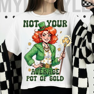 Może przedstawiać: Biała koszulka z grafiką rudowłosego skrzata trzymającego świecący kij z koniczyną. Tekst brzmi „NOT YOUR AVERAGE POT OF GOLD” w kolorze zielonym. Skrzat ma na sobie zielony kapelusz, kurtkę i złotą biżuterię.
