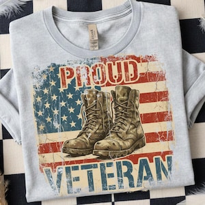 May include: Light gray t-shirt with a distressed American flag design. The flag graphic features combat boots and the words "PROUD VETERAN" in red and blue. The shirt is made of a soft material.