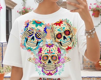 Puede incluir: Camiseta blanca con tres diseños de calaveras de azúcar coloridas. Las calaveras tienen intrincados patrones florales en azul, rojo, amarillo y morado. Una calavera se cubre los ojos, otra los oídos y la tercera la boca con huesos.