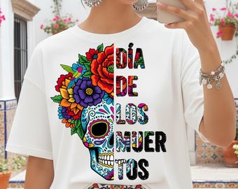 Puede incluir: Camiseta blanca con un diseño de calavera de azúcar colorida y acentos florales, con el texto "Día de los Muertos". El diseño utiliza colores vibrantes como el rojo, naranja, morado y azul, celebrando el Día de los Muertos.