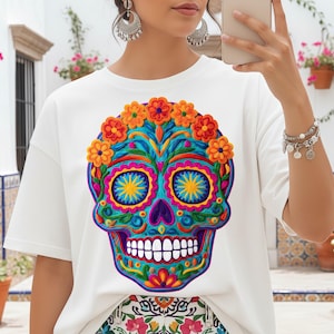 Puede incluir: Camiseta blanca con un diseño vibrante y colorido de calavera de azúcar. La calavera está decorada con intrincados patrones florales en tonos de azul, naranja, amarillo y morado. El diseño está centrado en la camiseta, creando un look llamativo.