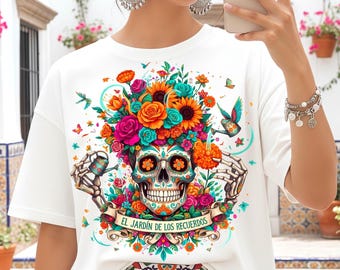 Puede incluir: Camiseta blanca con un diseño de calavera de azúcar colorido con elementos florales, incluyendo rosas y girasoles. La calavera tiene cuencas de ojos decorativas y el texto "EL JARDÍN DE LOS RECUERDOS" en una pancarta.