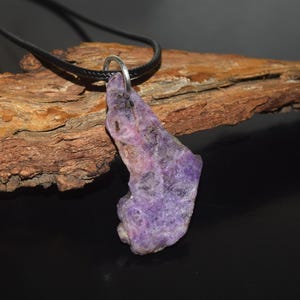 May include: A raw, purple amethyst pendant hangs from a black cord necklace. The irregularly shaped stone has a rough texture and varying shades of purple. The necklace is displayed on a piece of wood against a dark background.
