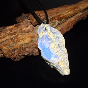 May include: A pendant necklace featuring a large, irregularly shaped stone with a light blue and beige color scheme. The stone is suspended from a black cord. The background includes a piece of wood and a black surface.