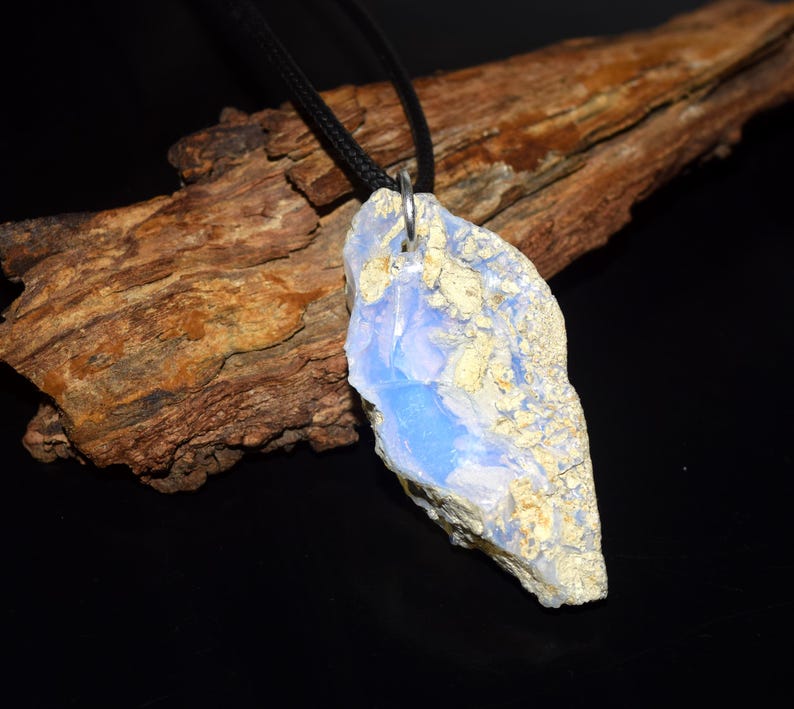 May include: A pendant necklace featuring a large, irregularly shaped stone with a light blue and beige color scheme. The stone is suspended from a black cord. The background includes a piece of wood and a black surface.