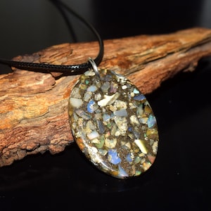 May include: An oval-shaped pendant featuring a mosaic of small, multicolored iridescent stone fragments embedded in a dark, textured resin base. The pendant is attached to a thin black cord, creating a necklace with a polished, smooth surface.